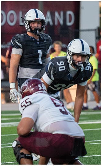 Two people wearing football helmets and dark jerseys are on a field with a blurred background showing part of a sign and some spectators. Descripci n generada con IA