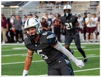 Two people in football uniforms are actively engaged on a field with one person crouched low and the other running toward them. Descripci n generada con IA