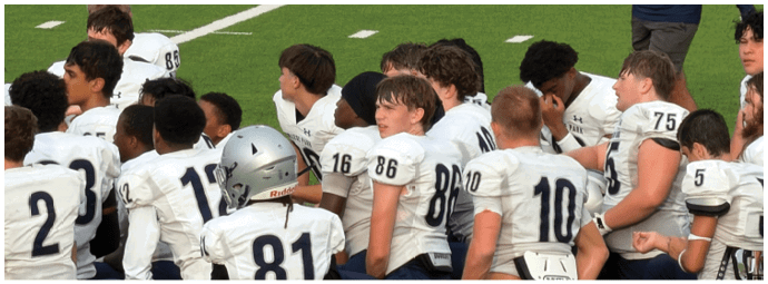 Five people wearing football helmets and uniforms stand on a field facing away from the camera with a group of people in the background. Descripci n generada con IA