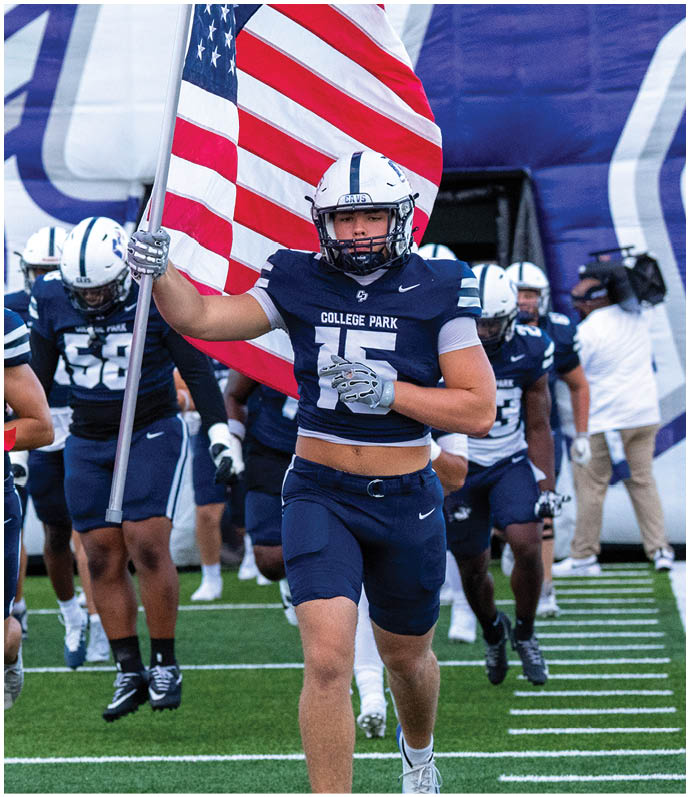 A person wearing a football helmet and uniform is holding a large American flag. Descripci n generada con IA