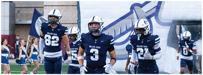 Four people in dark blue football uniforms with white helmets walk on a field with cheerleaders and a large inflatable tunnel in the background. Descripci n generada con IA