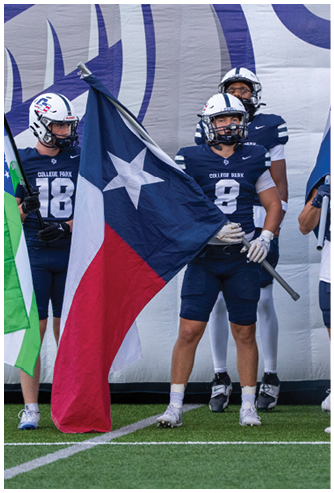 Three people in football uniforms stand on a field, with one holding a large Texas flag. Descripci n generada con IA
