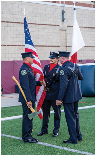 Three people in formal uniform stand together holding flags, including an American flag, in an outdoor setting. Descripci n generada con IA
