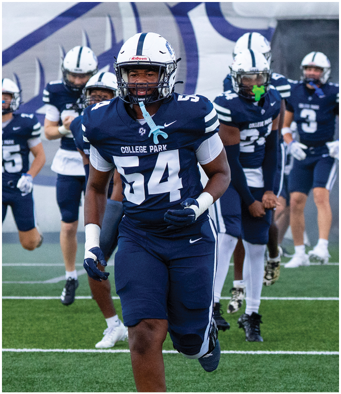 A person wearing a blue and white football uniform with the number 54 and a helmet is standing in front of other similarly dressed people on a field. Descripci n generada con IA