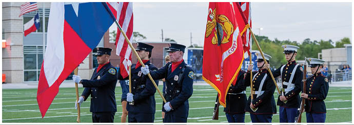 People in uniform stand in formation holding flags on a sports field with a building and bleachers in the background. Descripci n generada con IA
