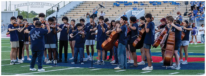 A group of people wearing matching dark blue shirts are playing string instruments in front of mostly empty bleachers at an outdoor venue. Descripci n generada con IA