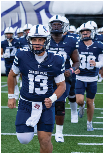Two people wearing blue College Park football uniforms and helmets are standing on a field, with one person in the foreground and the other slightly behind. Descripci n generada con IA