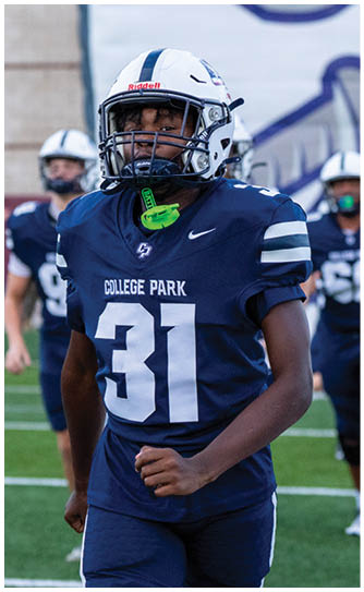 A person wearing a navy blue football uniform with the number 31 and a white helmet is standing on a football field with other players in the background. Descripci n generada con IA