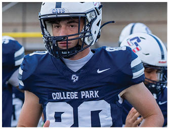 A person wearing a white football helmet with a blue stripe and the word “CAVS" on the front is on a football field with a scoreboard in the background. Descripci n generada con IA