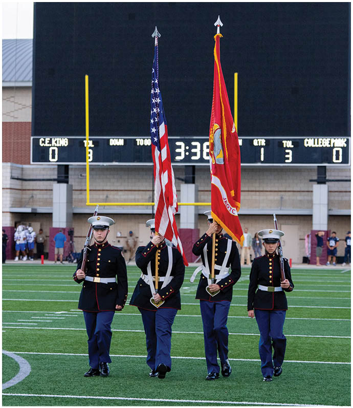 Four people in military dress uniforms stand on a football field holding rifles and two flags in front of a large scoreboard at Woodforest Bank Stadium. Descripci n generada con IA