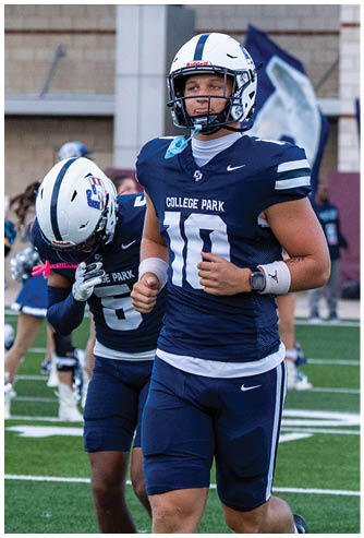 A person in a navy blue football uniform with the number 10 and a white helmet is running on a football field with another person in a similar uniform behind them. Descripci n generada con IA