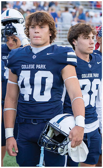 A person wearing a blue College Park football uniform with the number 40 is holding a white helmet and standing on a field with other players in similar uniforms. Descripci n generada con IA