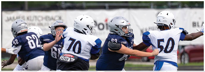 Four people wearing football helmets and white Bishop Verot jerseys stand side by side on a field with empty bleachers in the background. Descripci n generada con IA