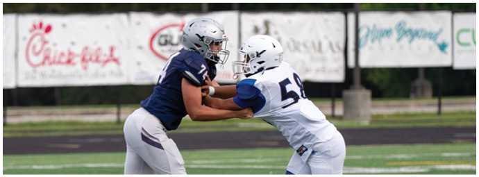 Four people wearing football helmets and white jerseys with “Bishop Verot" written on them stand side by side on a sports field with empty bleachers in the background. Descripci n generada con IA