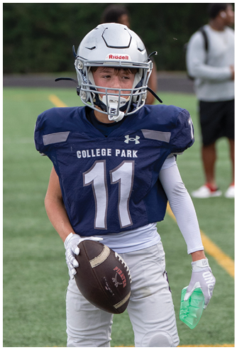 A person wearing a black helmet and a white football uniform with “Bishop Verot" on the jersey is running on a field with another player blurred in the background. Descripci n generada con IA