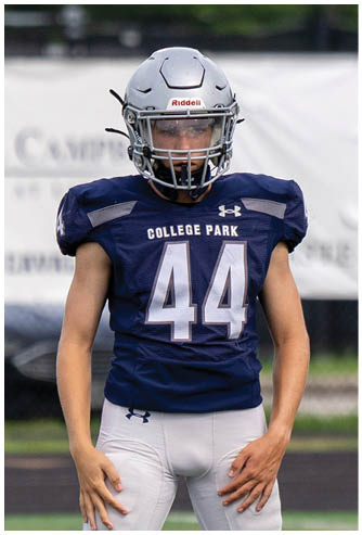 A person wearing a black football helmet and white Bishop Verot jersey with the number 66 stands on a football field with blurred players and bleachers in the background. Descripci n generada con IA
