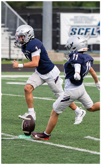 Two people in white and black football uniforms with helmets stand on a green field while other players and spectators are visible in the background. Descripci n generada con IA