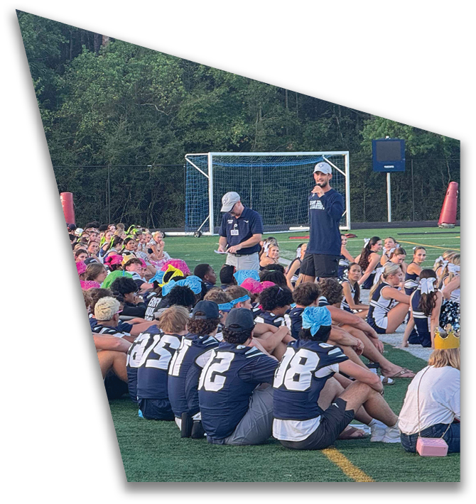 A large group of people wearing colorful clothing and headbands are gathered on a sports field while an adult in a cap and polo shirt stands nearby holding a clipboard. Descripci n generada con IA
