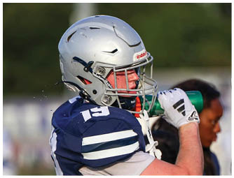 A person wearing a silver football helmet and navy blue jersey is drinking from a green water bottle on a sports field. Descripci n generada con IA