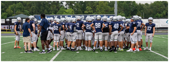 Four people wearing football helmets and white jerseys with “Bishop Verot" written on them stand side by side on a sports field with empty bleachers in the background. Descripci n generada con IA