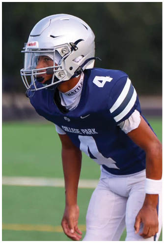 A person wearing a silver football helmet and navy blue jersey is drinking from a green water bottle on a sports field. Descripci n generada con IA