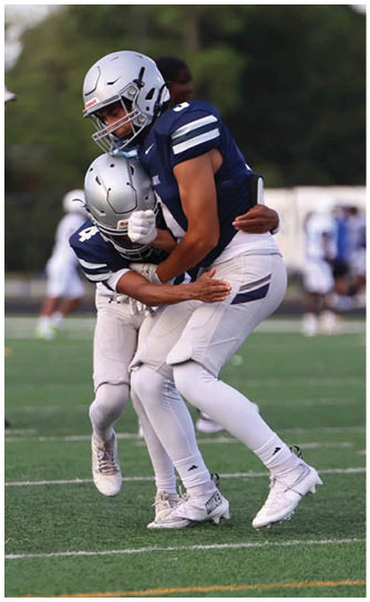 Two people wearing football uniforms and helmets are engaged in a tackle on a grassy field during a game. Descripci n generada con IA