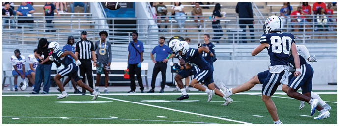 People in sports uniforms are playing football on a field with spectators sitting in bleachers in the background. Descripci n generada con IA