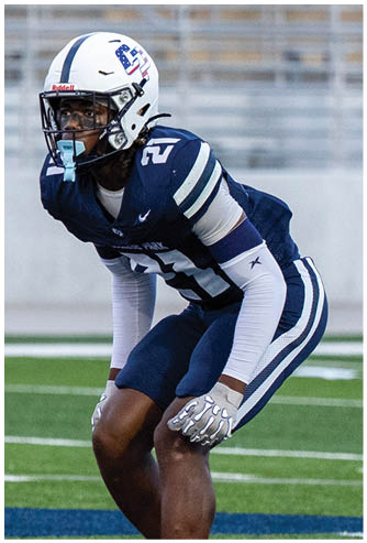 A person wearing a blue and white football uniform and helmet is crouched on a field, appearing focused and ready for action. Descripci n generada con IA