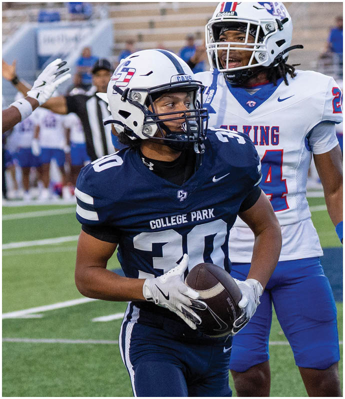 A person in a blue football uniform and helmet is on a field with another person reaching out a gloved hand in the background near a referee and blurred spectators. Descripci n generada con IA