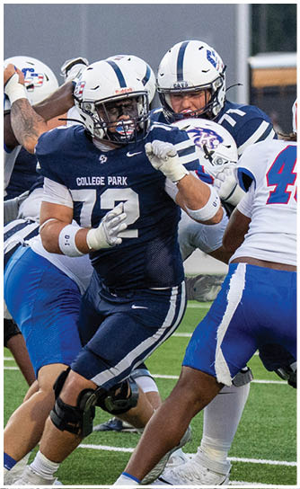 Several people in football uniforms are engaged in a play on a green field with one person in a dark uniform holding the ball while others in white and blue uniforms attempt to block or tackle. Descripci n generada con IA