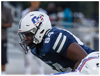 A person in a blue football uniform and white helmet with an American flag design is crouched in a ready position on a football field. Descripci n generada con IA