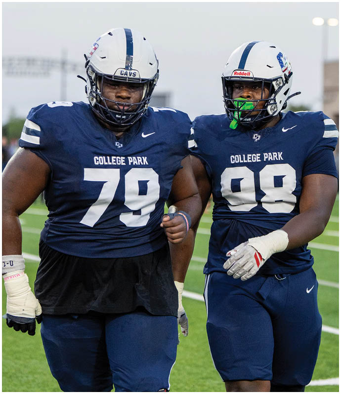 Two people wearing navy blue College Park football uniforms and white helmets stand side by side on a field. Descripci n generada con IA