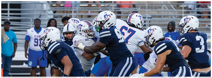 People in football uniforms are engaged in a play on a field with spectators and team members visible in the background. Descripci n generada con IA