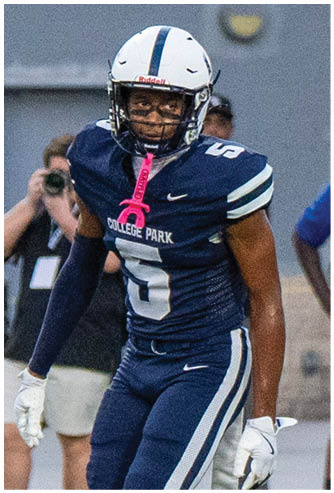 A person in a white and blue football uniform is lunging forward while being blocked by a person in a dark blue uniform during a football game on a green field. Descripci n generada con IA