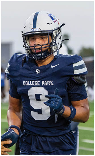 A person wearing a football helmet and navy blue jersey with “COLLEGE PARK" and the number 8 is on a field with blurred figures in the background. Descripci n generada con IA