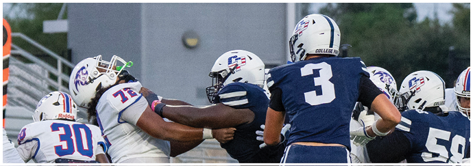 People wearing football uniforms are engaged in a play on a football field with one person in dark clothing being blocked by others in white and blue uniforms. Descripci n generada con IA