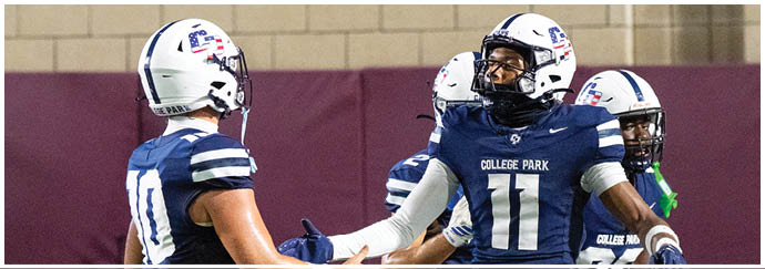 People wearing football uniforms are engaged in a play on a football field with one person in dark clothing being blocked by others in white and blue uniforms. Descripci n generada con IA