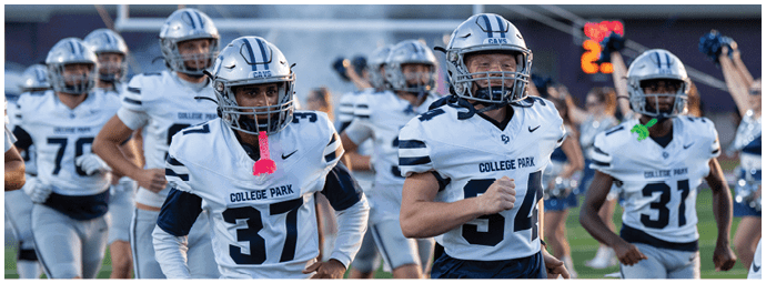 Four people in dark blue football uniforms with white helmets walk on a field with cheerleaders and a large inflatable tunnel in the background. Descripci n generada con IA