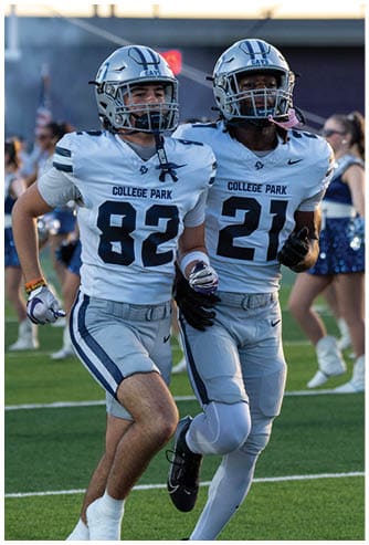 Two people wearing blue College Park football uniforms and helmets are standing on a field, with one person in the foreground and the other slightly behind. Descripci n generada con IA