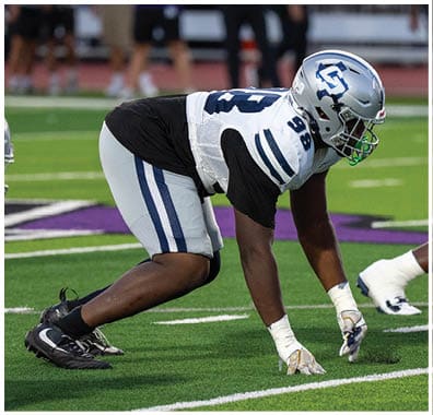A person wearing a black football uniform with the number one and a white helmet is on a football field with empty bleachers in the background. Descripci n generada con IA