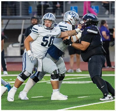 Two people in football uniforms are on a field with one holding a football and the other preparing to block or assist. Descripci n generada con IA