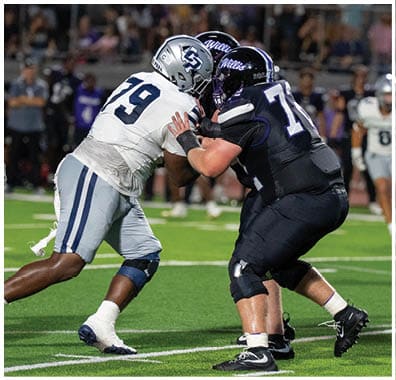A person wearing a black football uniform with the number three is giving a high five to a person in a white shirt and khaki pants on a football field during a game. Descripci n generada con IA