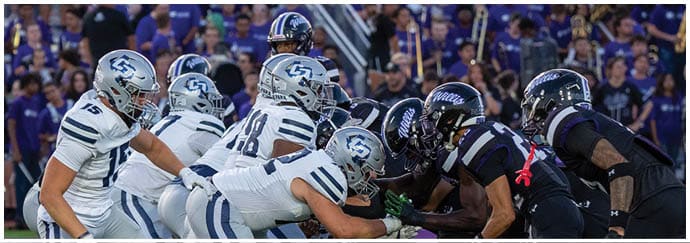 People wearing football uniforms are engaged in a play on a field with one person running while others attempt to block or tackle. Descripci n generada con IA