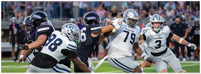 People wearing football uniforms are engaged in a play on a football field with one person in a white jersey blocking others in dark jerseys. Descripci n generada con IA