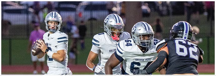 People wearing helmets and football uniforms are engaged in a close physical play on a football field with blurred spectators in the background. Descripci n generada con IA