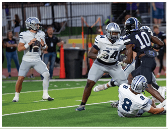A person in a dark sports uniform and helmet is crouched low on a football field near the sideline with several people standing in the background. Descripci n generada con IA