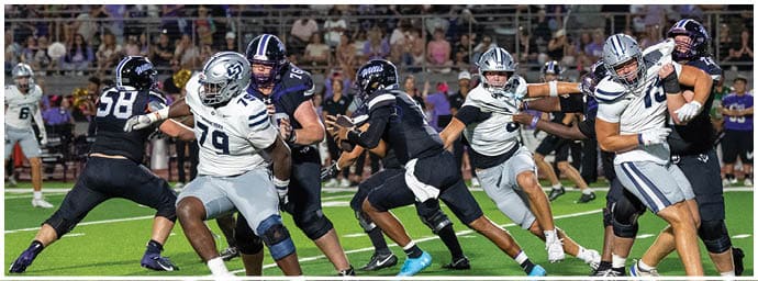 People wearing helmets and football uniforms are engaged in a play on a green football field with bleachers and spectators in the background. Descripci n generada con IA
