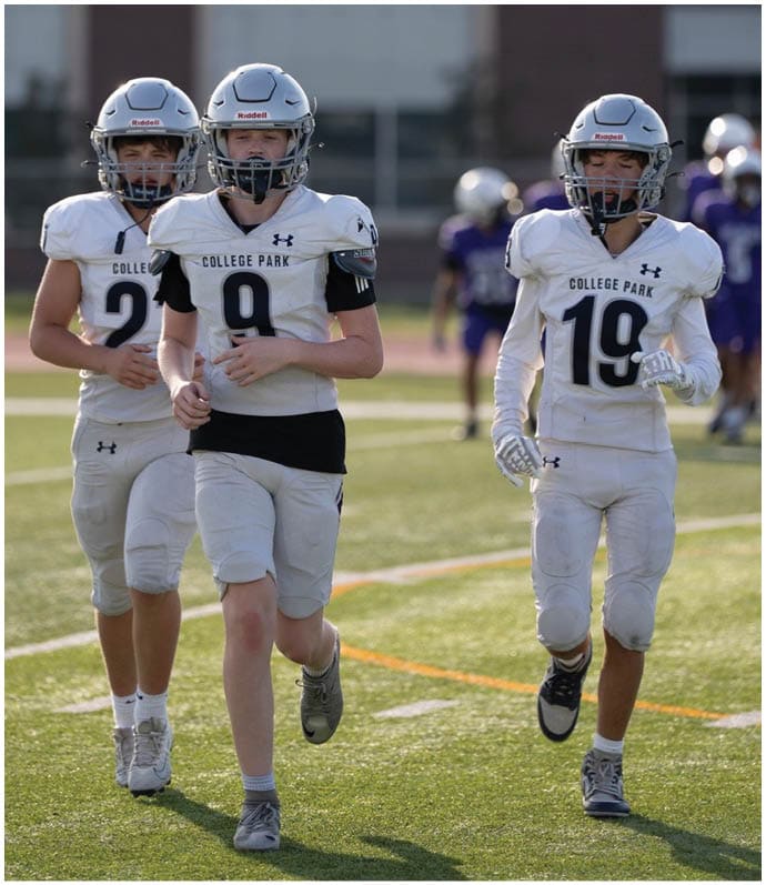 Three people wearing white football uniforms with helmets walk together on a green field during a game or practice. Descripci n generada con IA