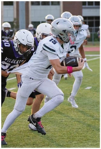 A person wearing a black football helmet and white Bishop Verot jersey with the number 66 stands on a football field with blurred players and bleachers in the background. Descripci n generada con IA