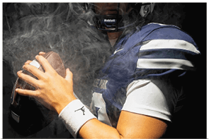 A person wearing a football helmet and uniform is holding a football with both hands surrounded by smoke. Descripci n generada con IA